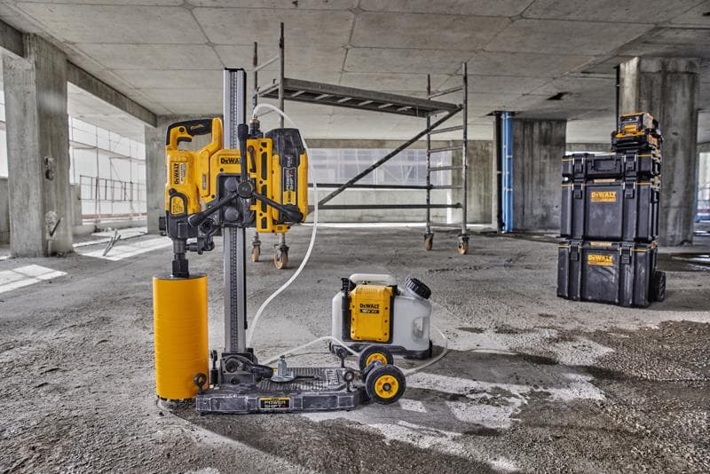 Een DEWALT kernboormachine met een gele cilindrische boor en aangesloten watertank staat op de vloer van een betonnen bouwplaats. Op de achtergrond zijn steigers en stapelbare opbergboxen van DEWALT te zien.