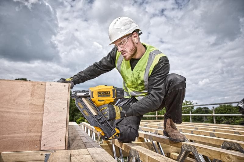 Persoon gebruikt een DEWALT nagelpistool DCN930N op houten planken, buiten op een bouwplaats met veiligheidsgerei.