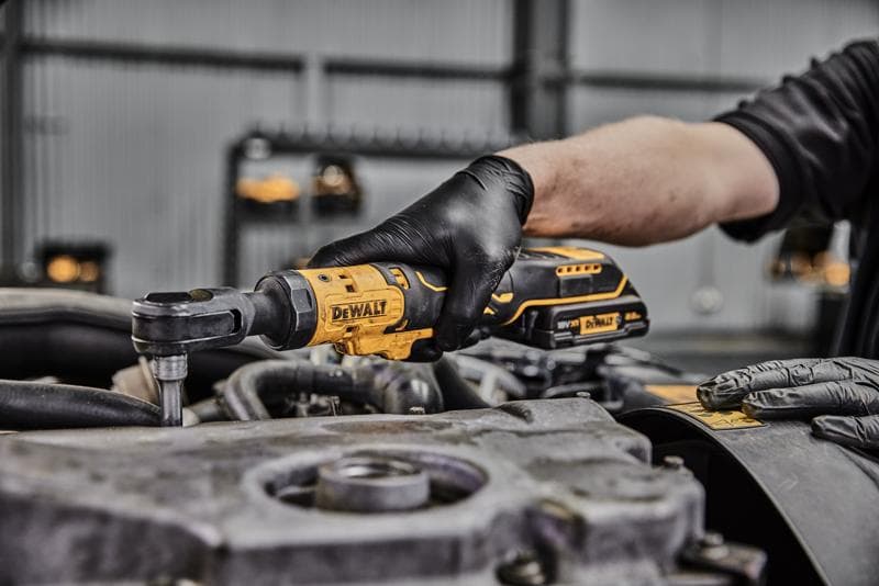 Een persoon met zwarte handschoenen gebruikt een DEWALT draadloze ratelsleutel bij een motoronderdeel in een werkplaats.