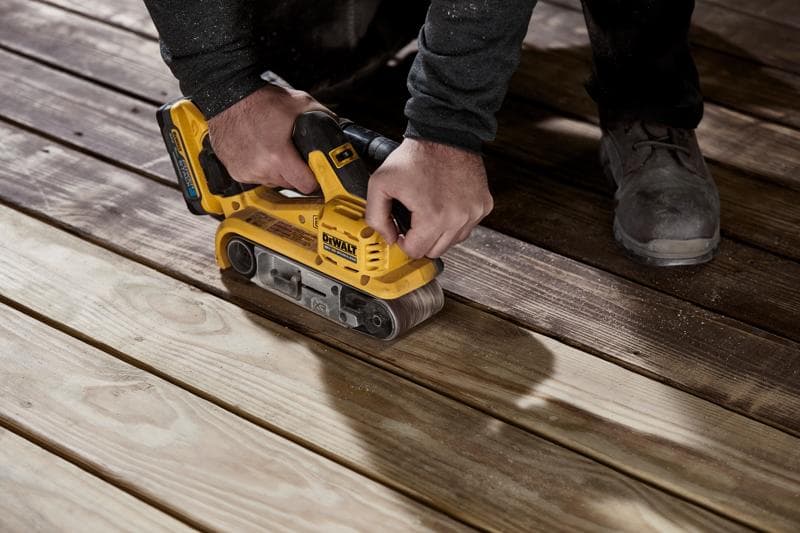 Een persoon gebruikt een gele DEWALT draadloze bandschuurmachine op houten planken om het oppervlak glad te maken.