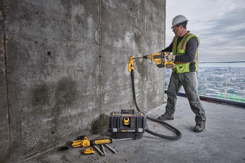 Een bouwvakker met een veiligheidsvest en helm gebruikt een draadloze DEWALT-boormachine met een stofafzuigaccessoire om een betonnen muur te boren. De werknemer staat op een groot gebouw met een uitzicht op de stad op de achtergrond. Verschillende DEWALT-gereedschappen en een gereedschapskoffer liggen nabij op de grond.