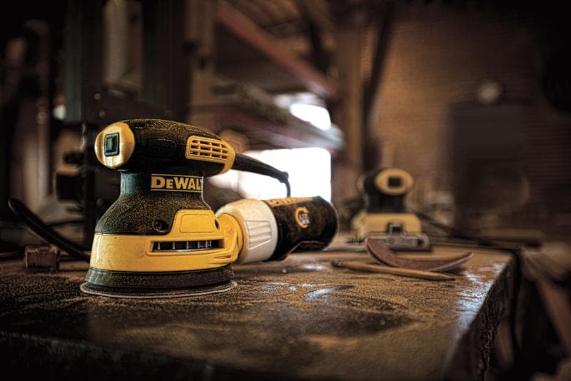 A close-up of a DEWALT DWE6421 electric sander on a dusty workbench in a workshop, with sanding pads and another sander visible in the background.