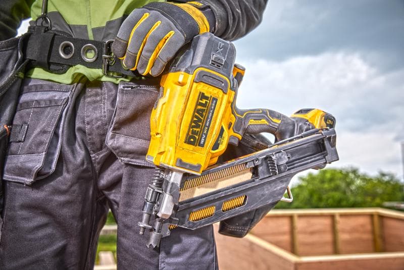 Persoon met een DEWALT DCN930N nagelpistool, handschoenen en werkuitrusting bij een bouwplaats buiten.