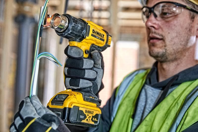Een persoon met werkhandschoenen en een veiligheidsvest gebruikt een DEWALT DCE530N draadloze heteluchtpistool om elektrische draden te verhitten op een bouwplaats. Het gezicht van de persoon is vervaagd voor privacy.