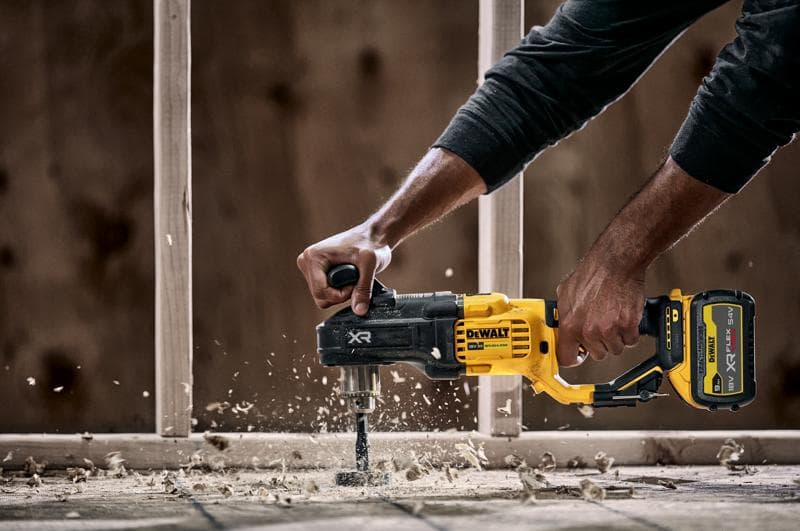 Een persoon gebruikt een DEWALT DCD444N draadloze boormachine met zijhandgreep om door hout te boren, wat houtspaanders en stof veroorzaakt. In de achtergrond is een bouwconstructie zichtbaar.