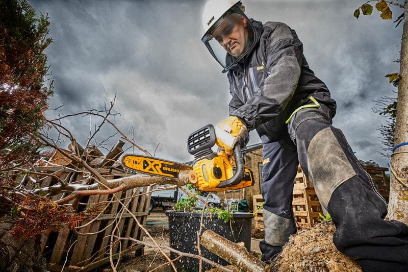 Persoon met veiligheidsuitrusting zaagt takken met een DEWALT kettingzaag buiten op een bewolkte dag.