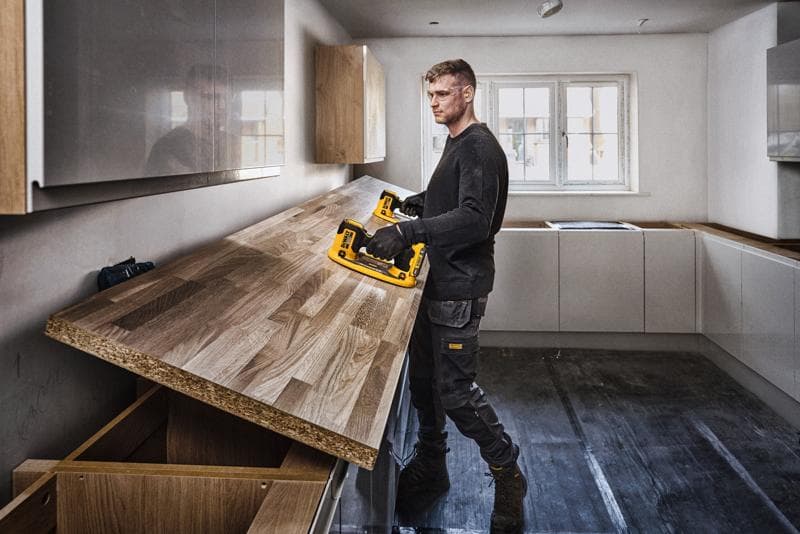 A person with a blurred face is installing a wooden countertop in a kitchen using a DEWALT power tool. The kitchen is modern with light-colored cabinets and a large window in the background.