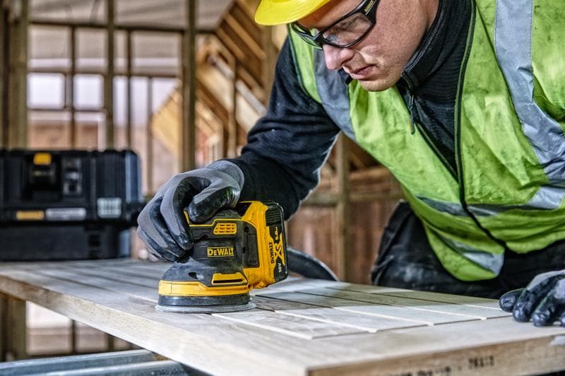 DEWALT DCW210, DCW210D1 schuurmachine gebruikt op hout, bouwlocatie, persoon draagt handschoenen en veiligheidsvest.
