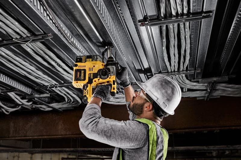 A person wearing a hard hat, safety glasses, gloves, and a safety vest uses a yellow DEWALT cordless rotary hammer drill to drill into a metal ceiling with electrical wiring.