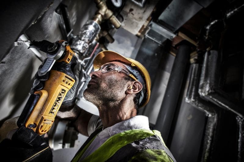 Werknemer gebruikt DEWALT DWE315 gereedschap om een pijp te snijden, draagt helm en vest op werkplek.