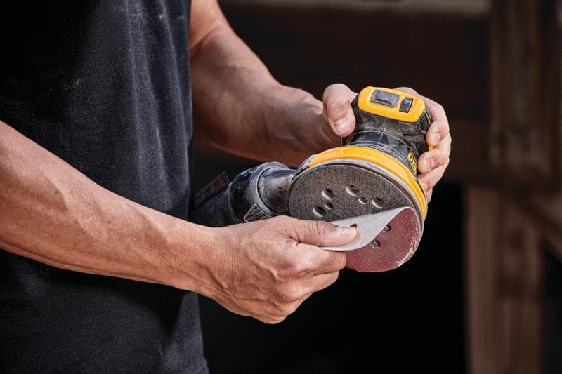 Een persoon houdt een DEWALT snoerloze excentrische schuurmachine, model DCW210B, vast en bevestigt een schuurpad aan het gereedschap.