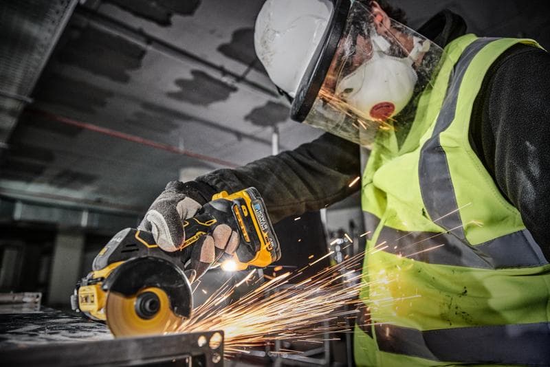 Een persoon met veiligheidsuitrusting, waaronder een helm, gezichtsscherm, masker, handschoenen en een reflecterend vest, gebruikt een DEWALT DCS438N draadloos gereedschap om metaal te snijden, wat vonken veroorzaakt.