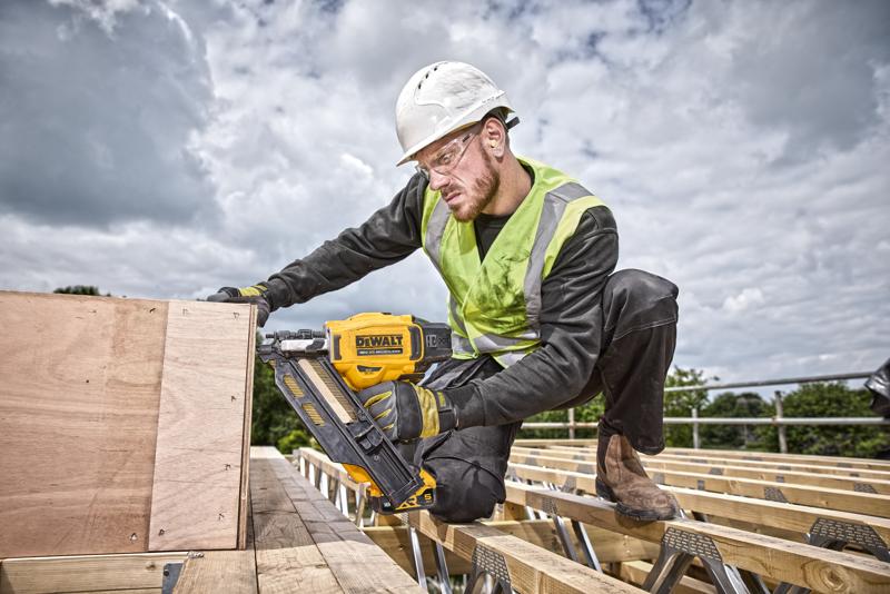 Persoon gebruikt een DEWALT nagelpistool DCN930N op houten planken, buiten op een bouwplaats met veiligheidsgerei.