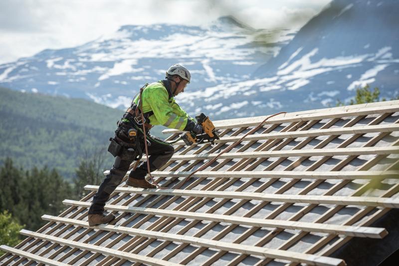 Werker gebruikt een DEWALT nagelmachine op een schuin dak buiten, met veiligheidsuitrusting en bergen op de achtergrond.