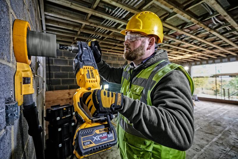 Een bouwvakker met een gele helm en een veiligheidshesje gebruikt een DEWALT DCD470N accuboormachine met een gatenzaag om in een betonnen muur op een bouwplaats te boren.