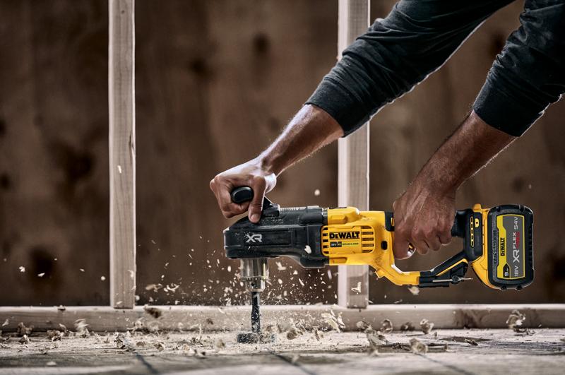 Een persoon gebruikt een DEWALT DCD444N draadloze boormachine met zijhandgreep om door hout te boren, wat houtspaanders en stof veroorzaakt. In de achtergrond is een bouwconstructie zichtbaar.