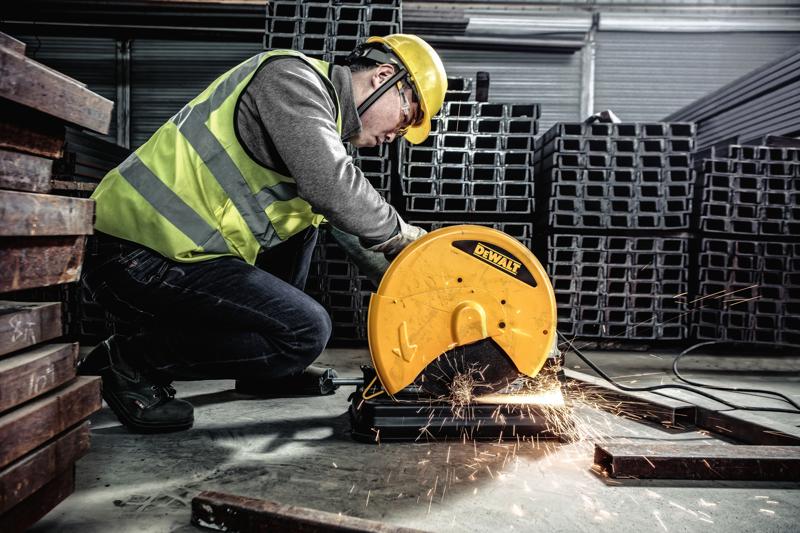 Een persoon met veiligheidskleding bedient een DEWALT metaalzaag, model D28730, om metalen staven te zagen in een industriële omgeving. Er komen vonken van het snijgebied.