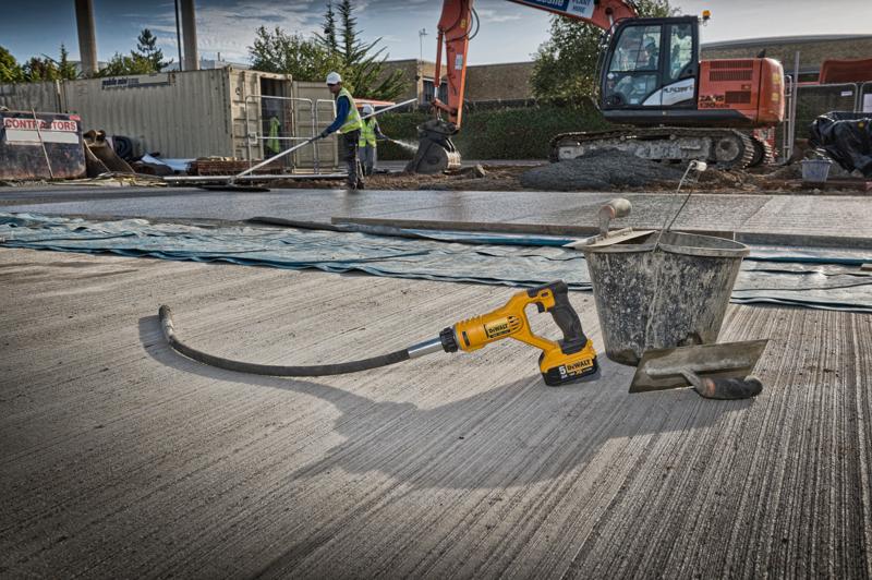 Een DEWALT DCE531N betontrilstok ligt op een bouwplaats naast een metalen emmer en een troffel op een vers gestorte betonnen ondergrond. Op de achtergrond zijn bouwvakkers, een grote graafmachine en andere bouwmaterialen te zien.