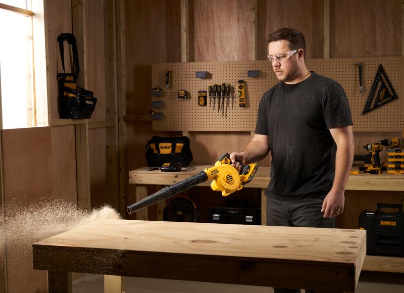 Persoon gebruikt een DEWALT DCV100 blazer om zaagsel van een houten werkbank te blazen in een werkplaats.