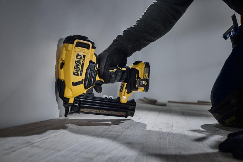A person using a DEWALT DCN210 cordless yellow and black nail gun to attach wooden trim along the bottom of a wall.